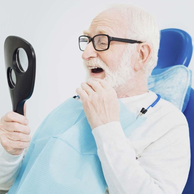 Älterer Herr im Zahnarztstuhl betrachtet seine Zähne im Spiegel